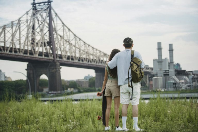 Back view of unrecognizable young male tourist in casual clothes with backpack cuddling girlfriend with skateboard in hand while standing on grassy ground near Brooklyn Bridge and admiring city